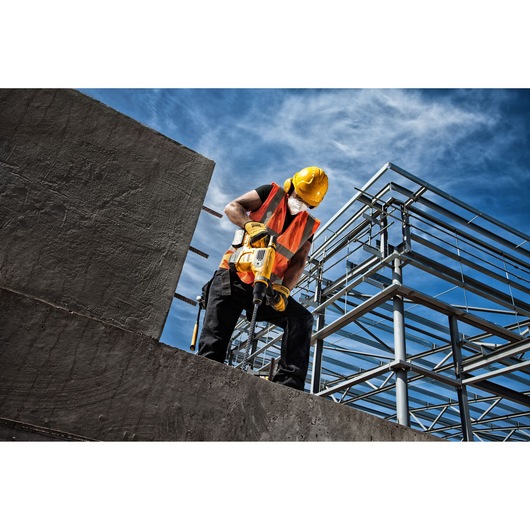 Worker in safety gear uses a yellow DEWALT drill on a concrete structure at a construction site with steel framework.
