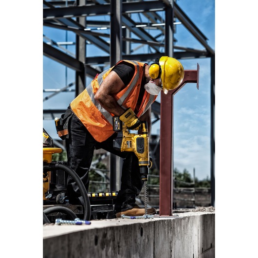 Worker using DEWALT drill on concrete at construction site, wearing safety gear and orange vest.