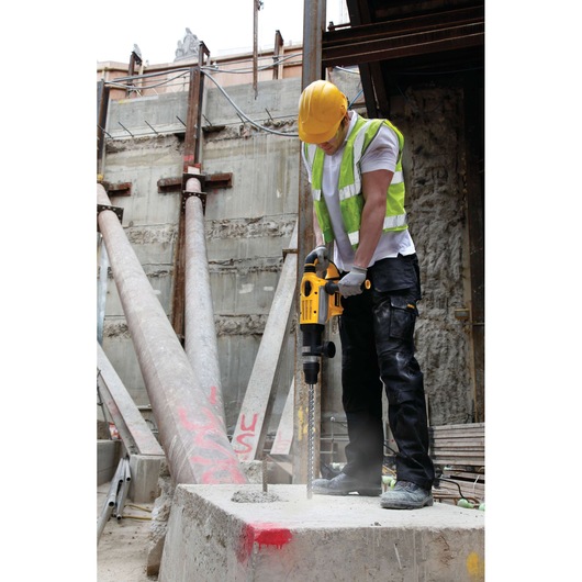 Worker uses DEWALT drill on concrete at a construction site, wearing a helmet and high-visibility vest.
