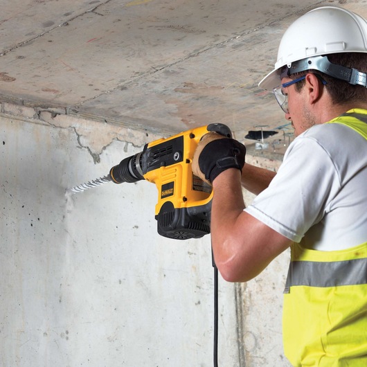 Worker drills concrete wall with DEWALT power tool, wearing safety gear and high-visibility vest.