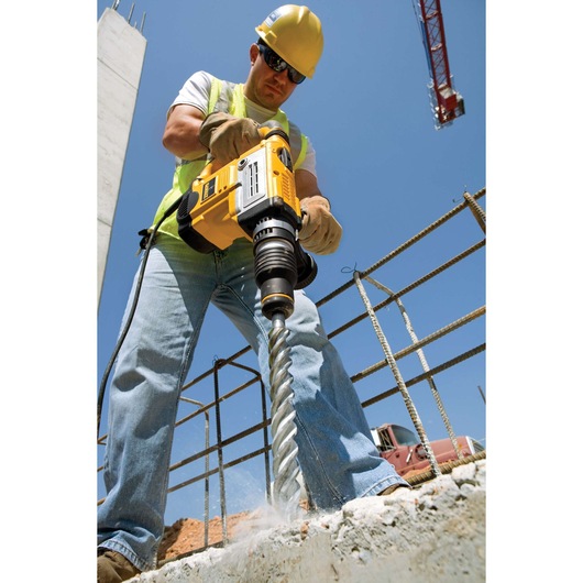 Worker uses a DEWALT drill with DT60844 bit on concrete at a construction site, wearing safety gear.