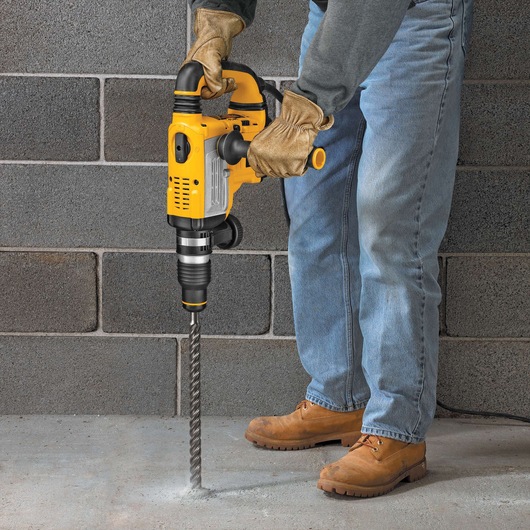 Person using a yellow DEWALT rotary hammer drill on concrete floor, wearing gloves and work boots.