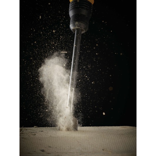 Chisel tool striking concrete surface, dust rising in dramatic effect against a black background.