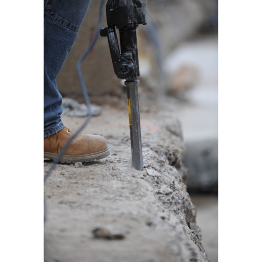 Worker uses a DEWALT chisel tool to break concrete on a construction site, wearing jeans and safety boots.