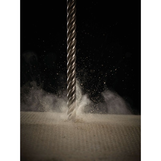 A metal spiral drill bit penetrates a surface, creating dust clouds against a black background.