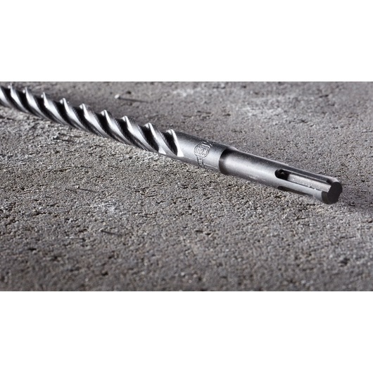A close-up image of a DEWALT SDS-Plus masonry drill bit lying on a rough concrete surface.