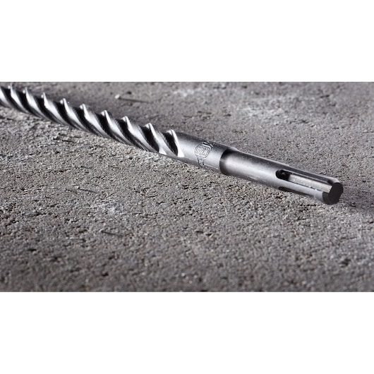 A close-up image of a DEWALT DT8973 SDS drill bit lying on a rough concrete surface. The drill bit features a spiral design for efficient drilling.