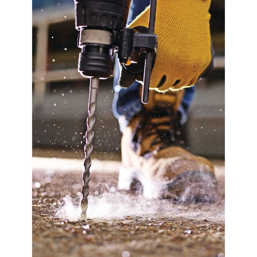 Gloved hand uses a DEWALT drill bit to drill into a concrete surface with dust and debris flying.