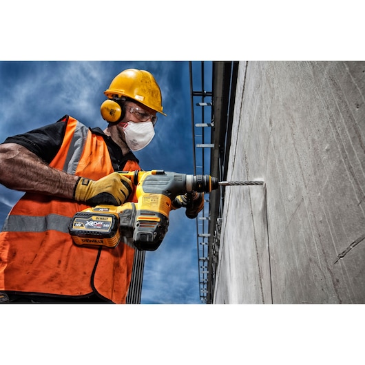 Worker in safety gear drills concrete wall with a DEWALT cordless rotary hammer under blue sky.