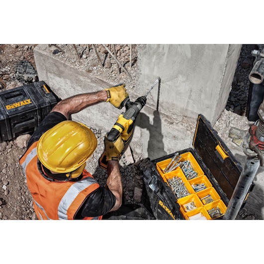 Worker using a DEWALT drill on concrete, with toolboxes and fasteners nearby on a construction site.