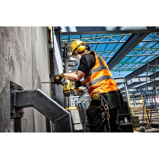 Worker uses a DEWALT drill on a concrete wall at a construction site, wearing safety gear and an orange vest.