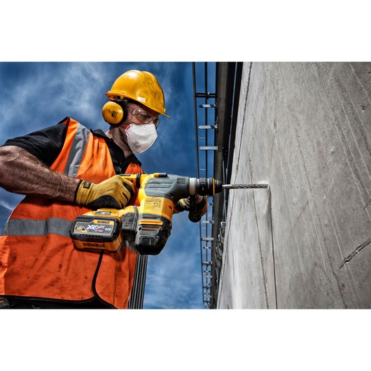 Worker using a DEWALT cordless drill with DT9609 bit to drill into a concrete wall outdoors, wearing safety gear.