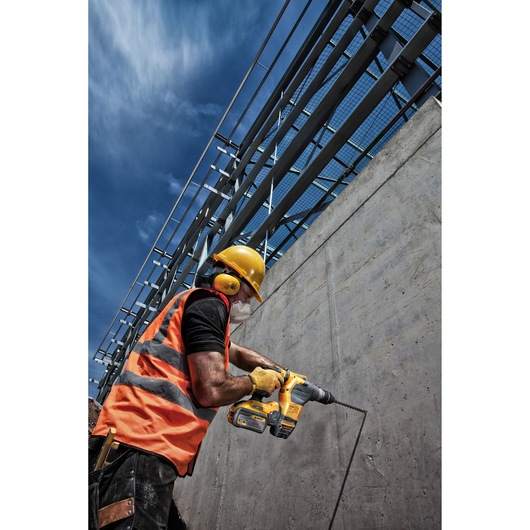 Worker in safety gear uses a DEWALT drill on a concrete wall at an outdoor construction site under a blue sky.