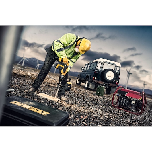 Worker uses DEWALT drill outdoors, wearing safety gear. SUV and wind turbines visible in background.