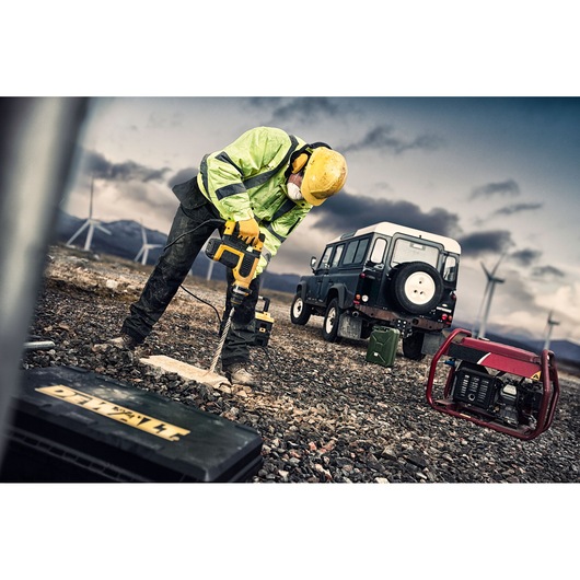 Worker in safety gear drills ground with DEWALT tool near SUV and generator, wind turbines in background.