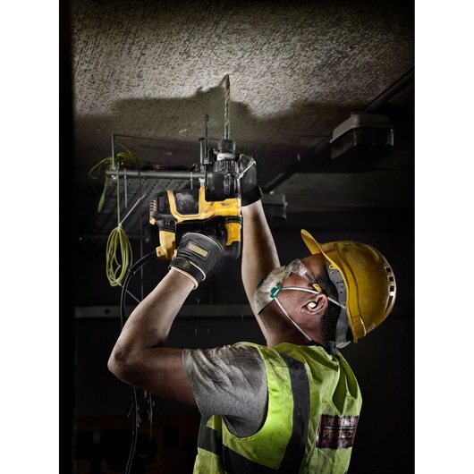 Worker drills ceiling with a yellow DEWALT tool, wearing safety gear and high-visibility vest.