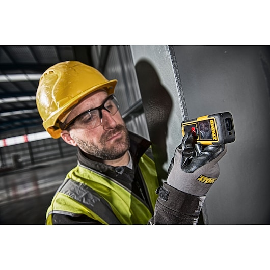 Person in safety gear using a DEWALT DW033 laser distance measurer in an industrial setting.