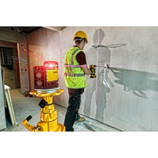 DEWALT laser level on tripod projects lines, worker uses drill in construction setting.