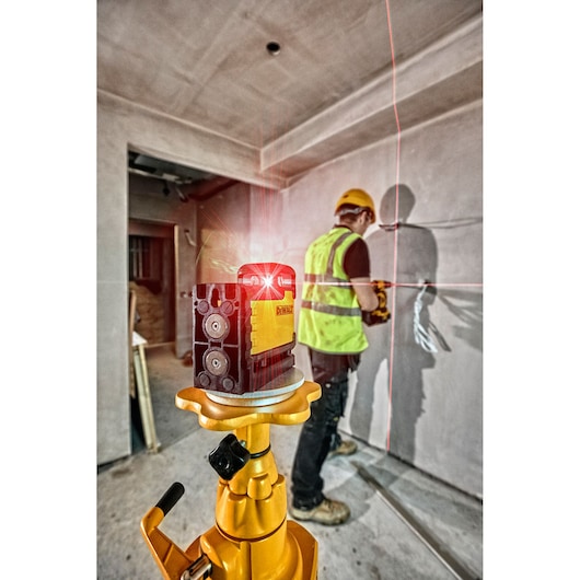 DEWALT laser level on tripod at a construction site, projecting red lines. Worker in safety gear is in the background.