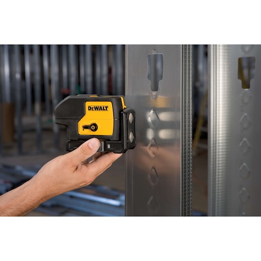 A person holding a DEWALT DW083K laser level tool against a metal framing stud at a construction site.