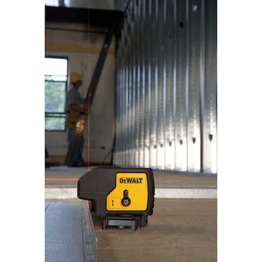 DEWALT laser level device model DW083K placed on a construction floor, projecting a red vertical line for alignment. Metallic wall frames and a construction worker in the background.