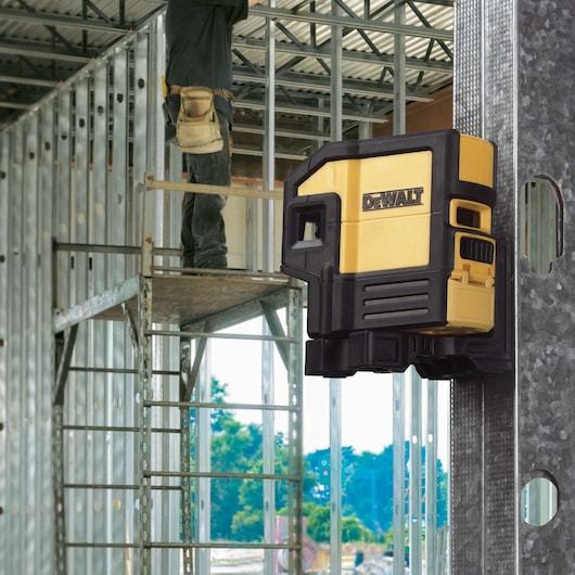 A DEWALT DW0851 yellow and black laser level device is mounted on a metal stud in a construction site, with a person working on scaffolding in the background.