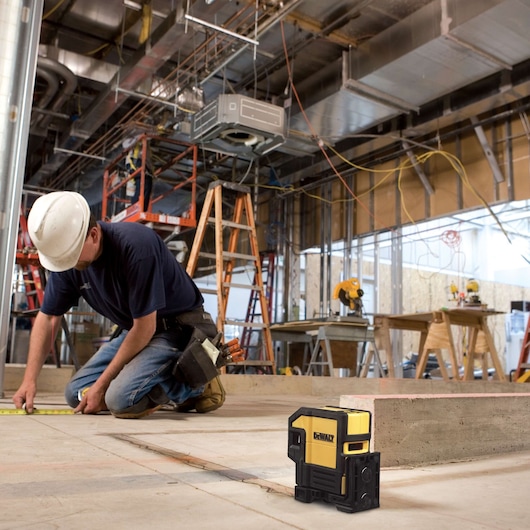A DEWALT DW0851 laser level is placed on the floor of a construction site. In the background, a worker wearing a hard hat is measuring the floor with a tape measure. There are ladders, scaffolding, and construction tools visible throughout the workspace.