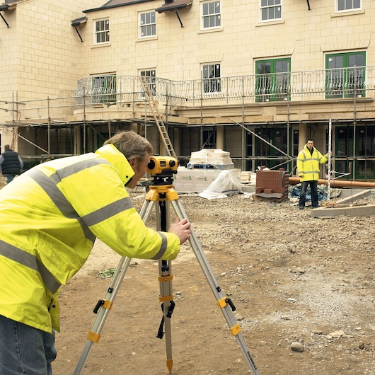 Worker uses DEWALT DW096PK level tripod on construction site, another holds measuring rod.