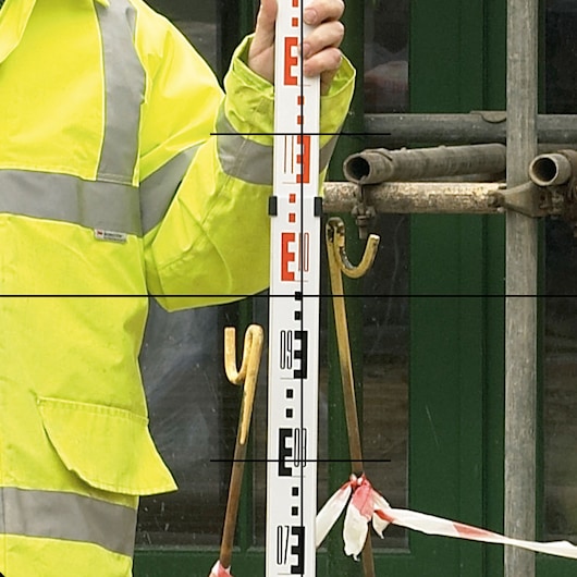 Person in high-visibility jacket holding a DEWALT leveling rod in front of construction equipment.