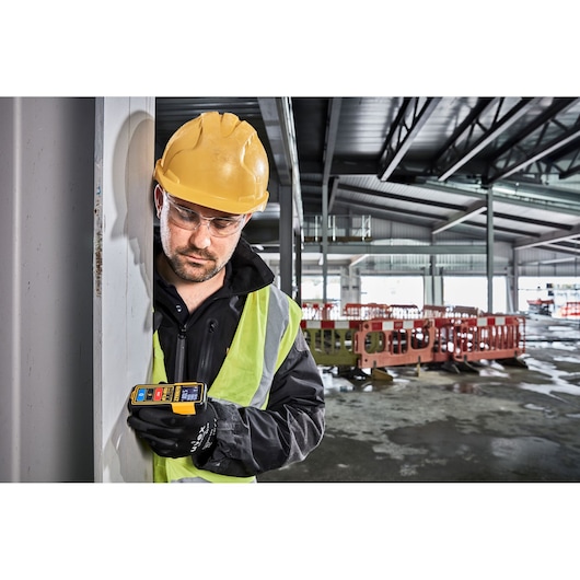 Worker in safety gear uses a DEWALT DW099S laser measure at a construction site indoors.