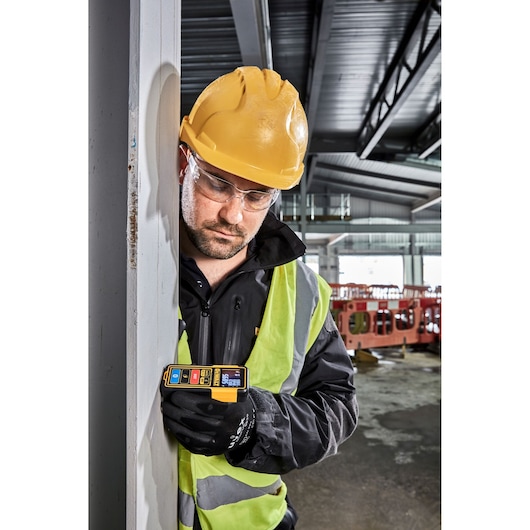Worker uses DEWALT DW099S laser distance measurer at a construction site, wearing safety vest and helmet.