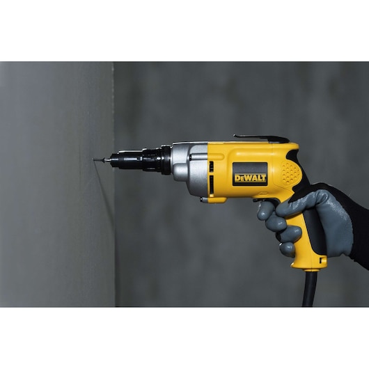 A person wearing a grey glove uses a yellow DEWALT electric screwdriver to drive a screw into a wall.