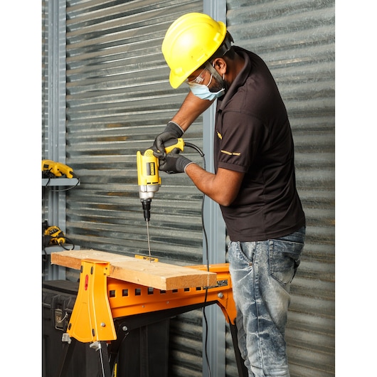 Person uses a DEWALT electric drill on wood at a workshop, wearing safety helmet and gloves.