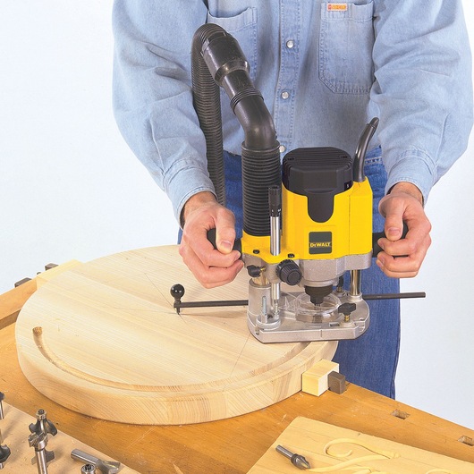 Person using a DEWALT DW622K router to cut a circular groove in a piece of wood on a workbench.