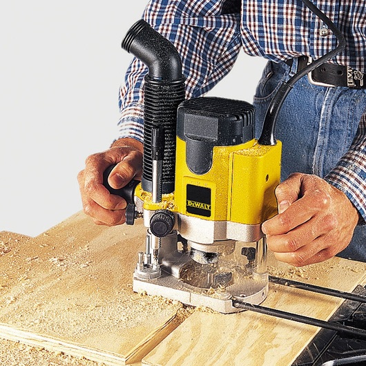 DEWALT DW622K router being used to cut wood, held by a person in plaid shirt, wood dust visible.
