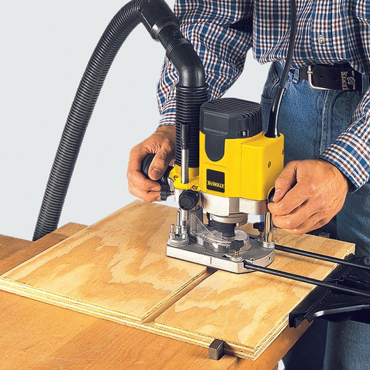 Person operating a DEWALT DW622K router, cutting wood with attached dust extraction hose.