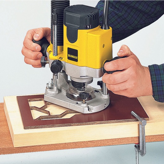 DEWALT DW622K router being used on wood with a stencil for precise shaping, held by hands.