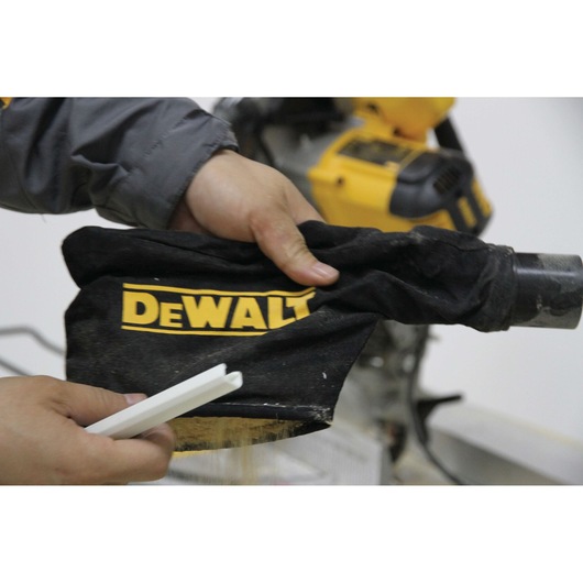 Hand holding DEWALT dust bag near yellow machine, used for collecting saw dust during woodworking.