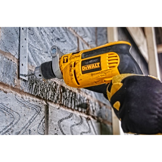 DEWALT drill in use, drilling into a metal bracket on a brick wall. Gloved hand holds the yellow power tool.