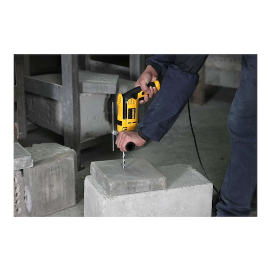 A person uses a yellow DEWALT electric drill to drill into a concrete block. The person is holding the drill with both hands for stability in an industrial setting.