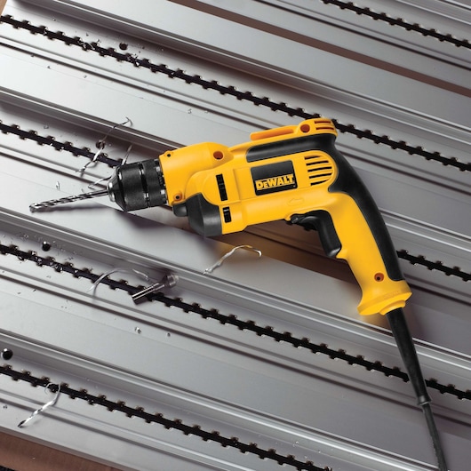 A yellow and black DEWALT corded electric drill with a metal drill bit, shown drilling into a silver metal surface with spiral metal shavings around the hole.