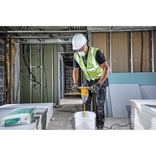 Worker uses a DEWALT DWD241 drill to mix materials in a bucket on a construction site.