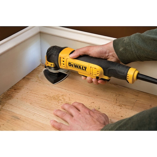 Hand using DEWALT oscillating multi-tool to sand wood floor near white baseboard corner.