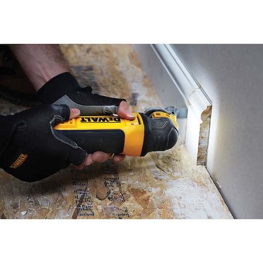 A DEWALT multi-tool cutting drywall near a baseboard, held by gloved hands on a wood floor.