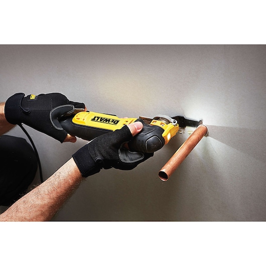 A person uses a DEWALT multi-tool to cut drywall around a copper pipe, wearing black safety gloves.