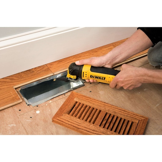 Person uses a DEWALT multi-tool to cut wood flooring near a floor vent cover.