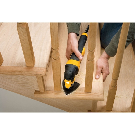 Person using a DEWALT multi-tool to sand wooden stairs, working between stair rails.