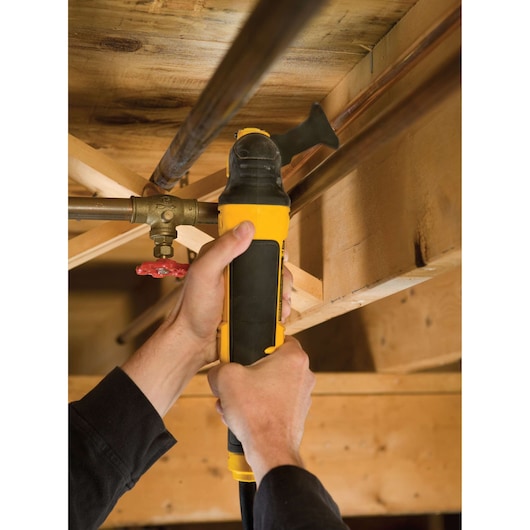 Hands using a DEWALT multi-tool on a pipe under wooden beams. Tool is yellow and black.