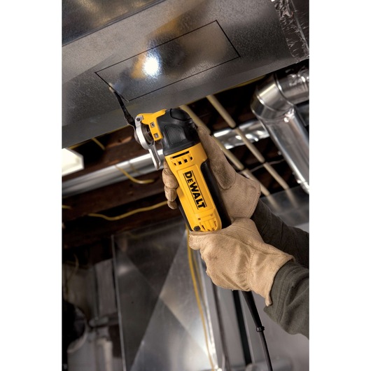 Gloved hands use a DEWALT oscillating multi-tool to cut a square in a metal duct with an attached work light.