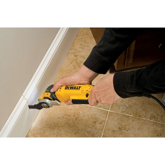 Hands using a DEWALT oscillating multi-tool to cut a white baseboard near a tiled floor.